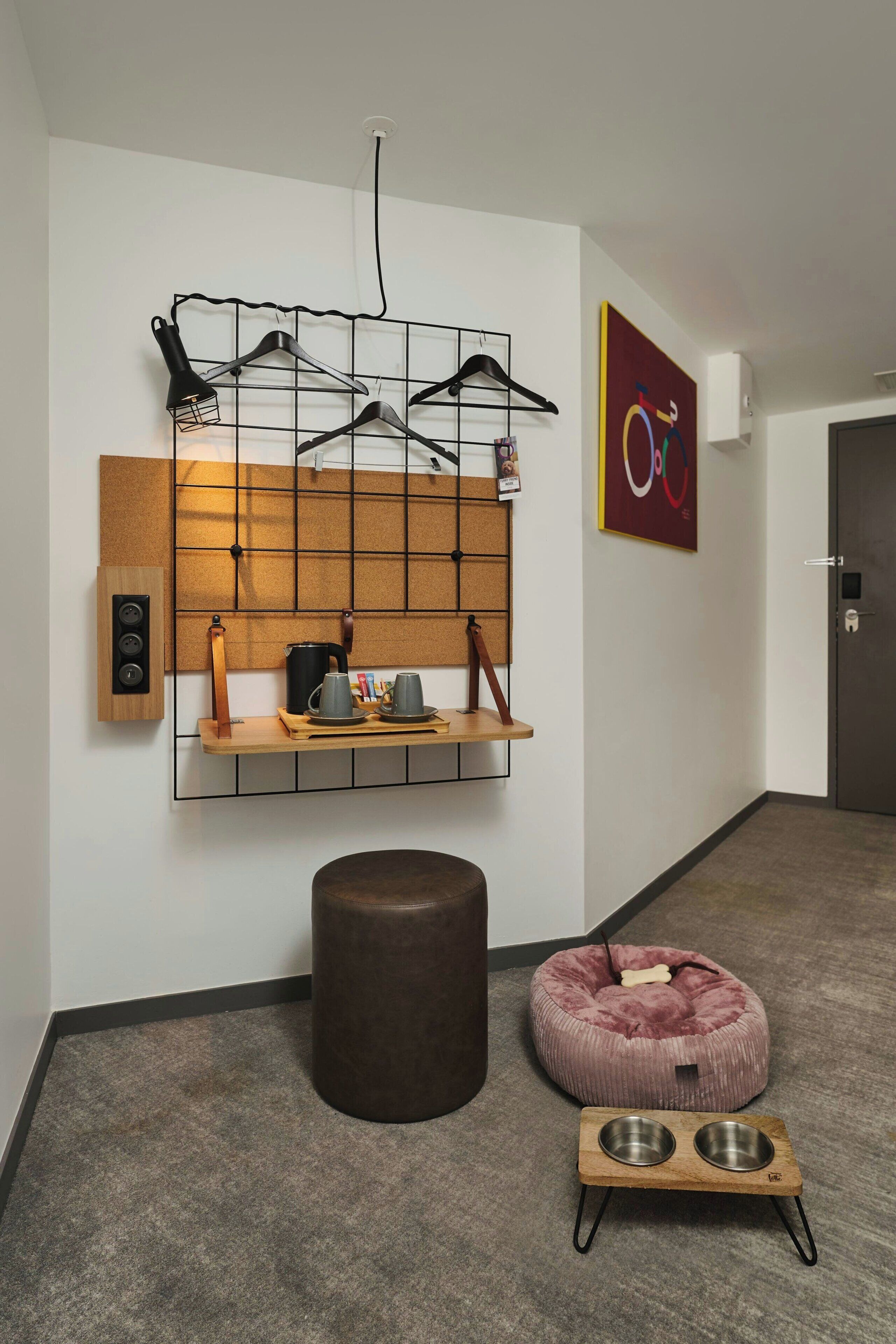 A modern interior with a wall-mounted rack holding black hangers, a wooden shelf with a kettle and mugs, a brown ottoman, a pink pet bed, and a metal pet bowl stand.