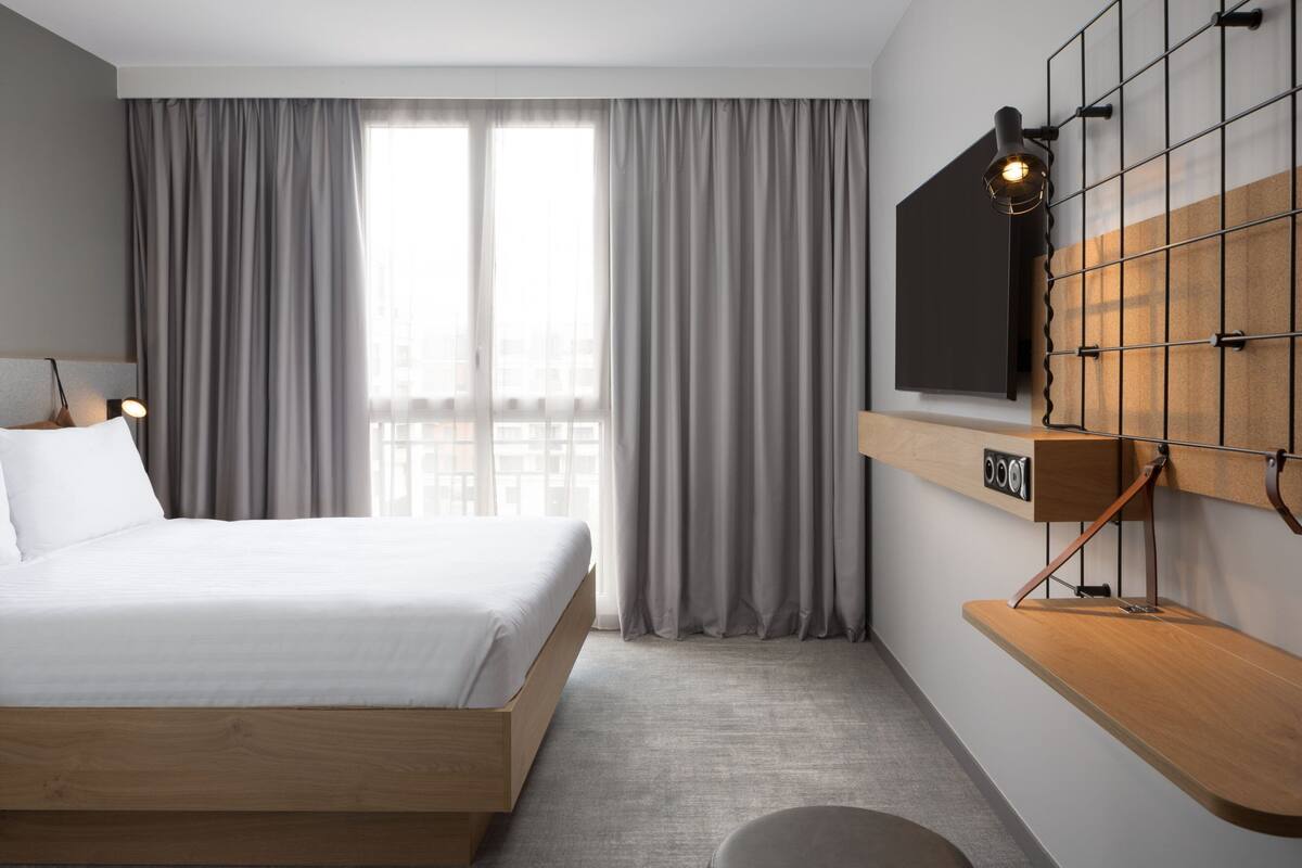 Quarto de hotel moderno com cama de madeira, cortinas cinzentas, televisão na parede e prateleira de madeira.