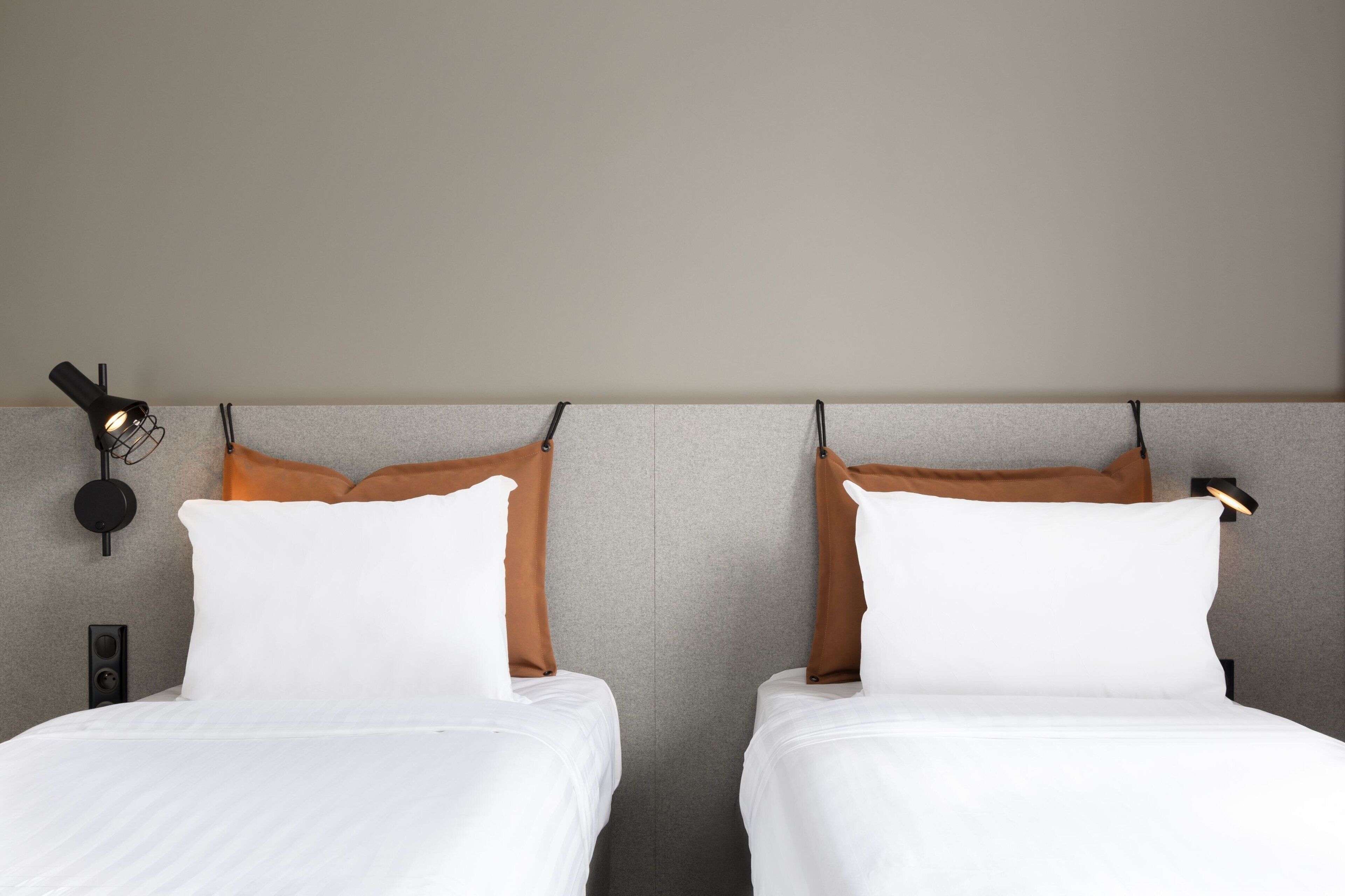 Two beds with white bedding and brown pillows, a wall-mounted light fixture, and a speaker on the left side of the bed.