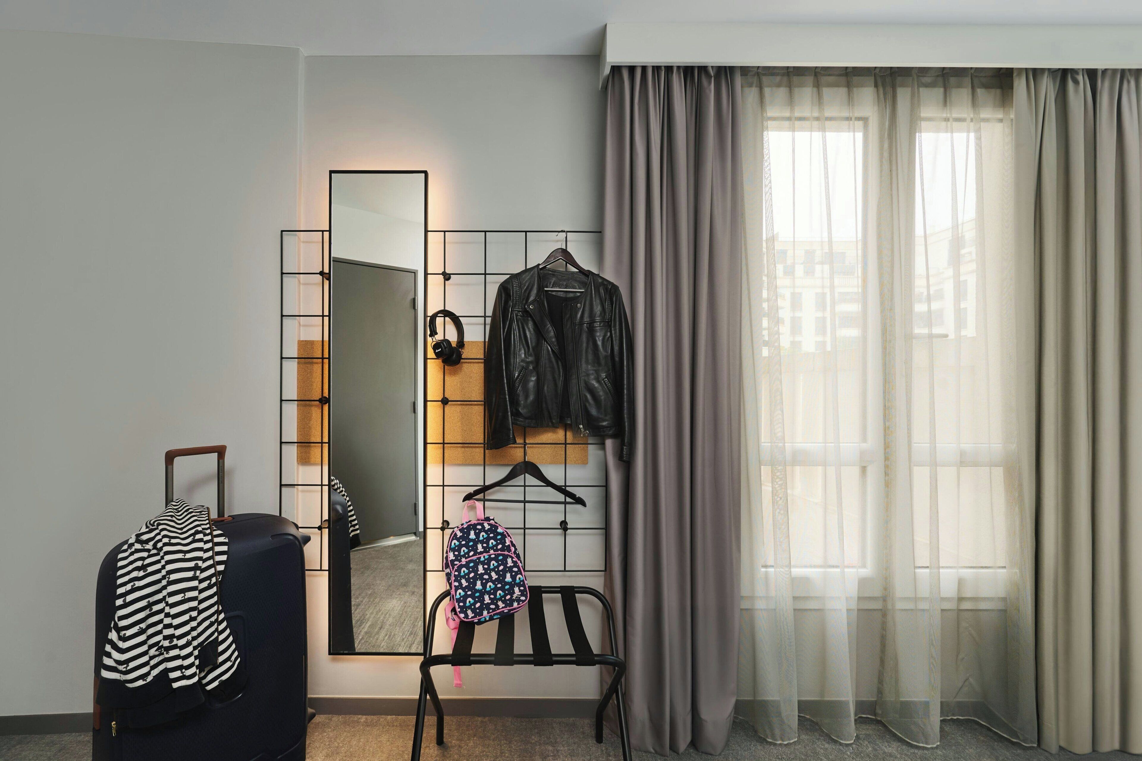A room with a large mirror, a black metal chair, a black suitcase, a backpack, and a leather jacket hanging on a rack.