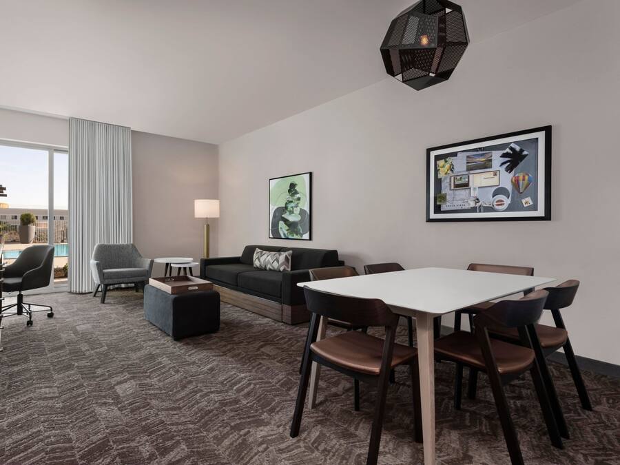 A modern hotel room with a large window, a sofa, a dining table, and a lamp.
