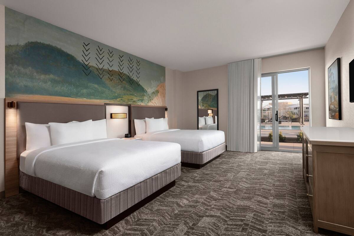 A hotel room with two beds, a large painting on the wall, a view through the glass door, and a desk with a lamp.