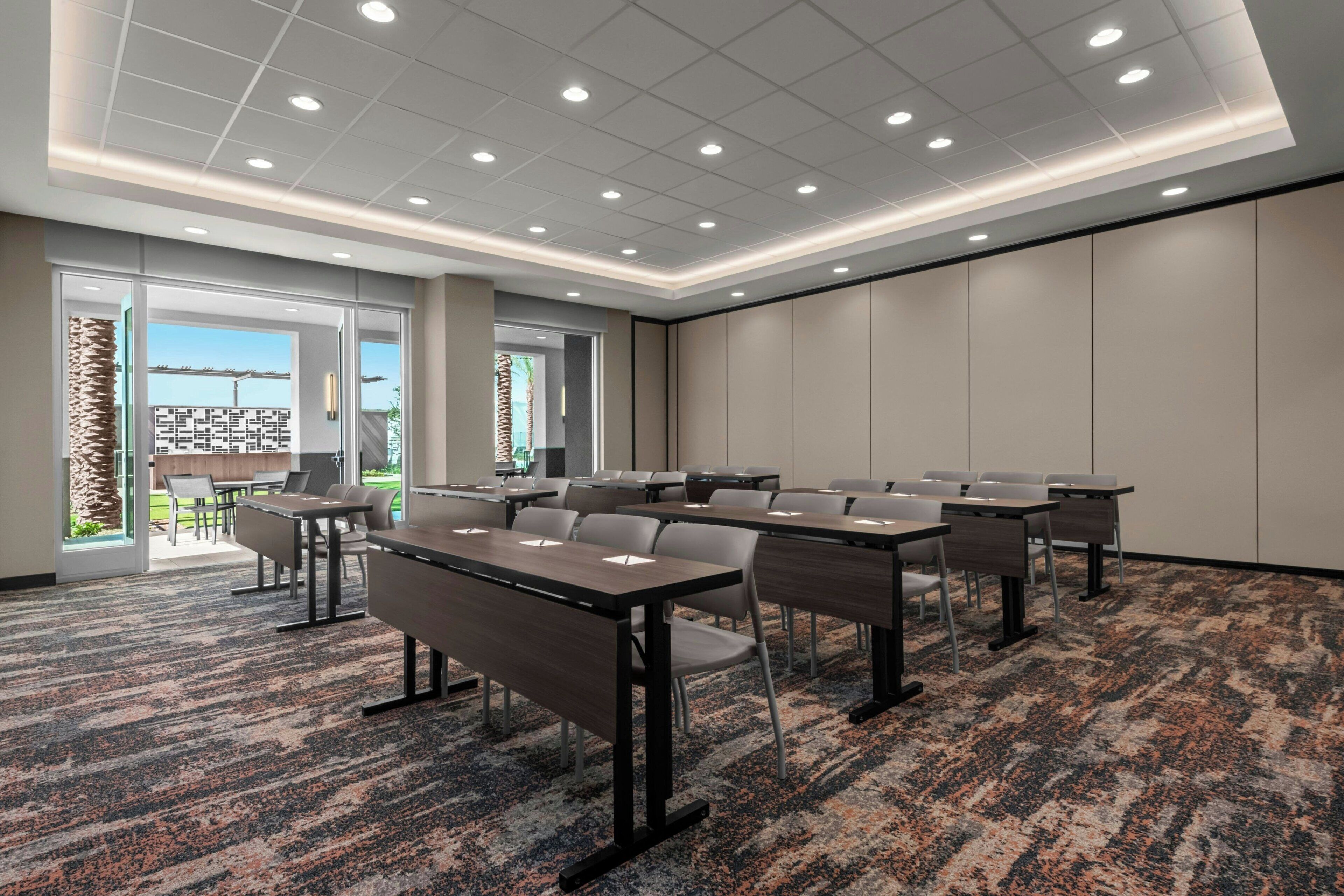 A conference room with multiple tables and chairs, large windows with a view of the outdoors, and a carpeted floor.