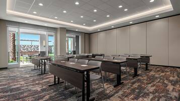 A conference room with multiple tables and chairs, large windows with a view of the outdoors, and a carpeted floor.