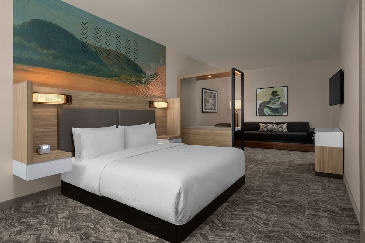 A modern hotel room with a large bed, a nightstand with a lamp, a sofa, and a painting on the wall.