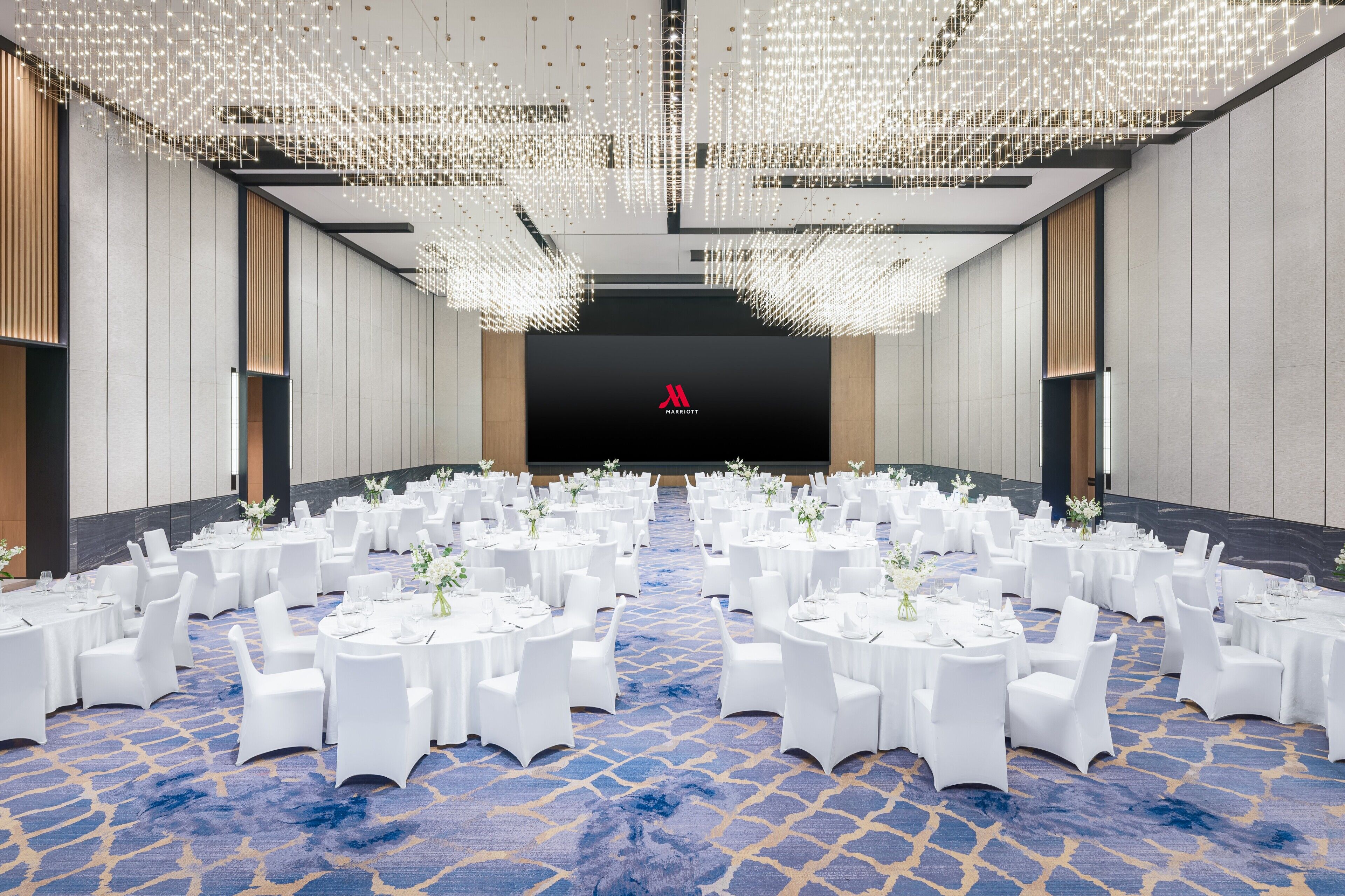 Un amplio salón de conferencias con mesas redondas y sillas blancas, un escenario con un logotipo rojo y un techo con luces colgantes.