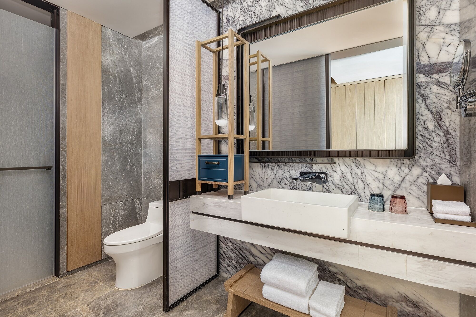 A modern bathroom with a marble wall, a wooden vanity, a white sink, and a toilet.