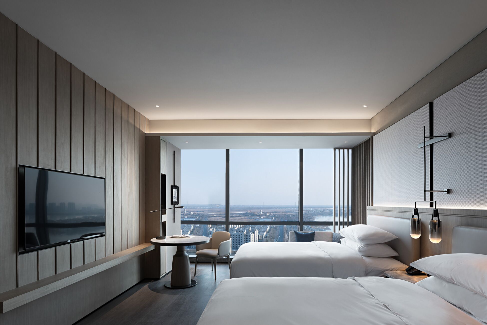 Habitación de hotel moderna con un televisor grande, una mesa redonda, dos camas y vista a la ciudad.
