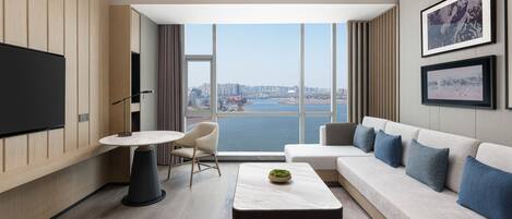 A modern hotel room with a large window, a flat-screen TV, a round table, a sofa, and a coffee table.