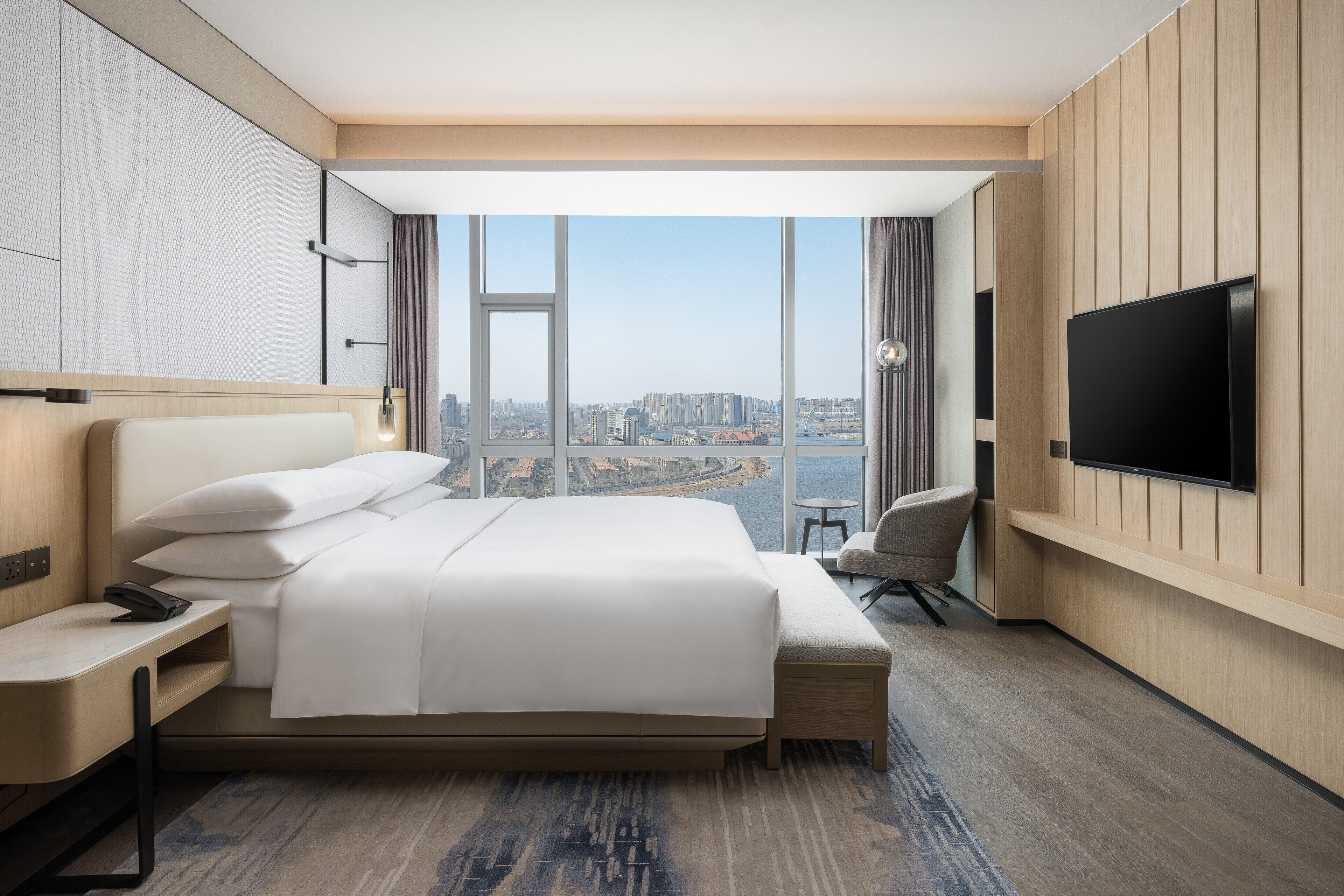 A modern hotel room with a large bed, a desk, a chair, a TV, and a city view.