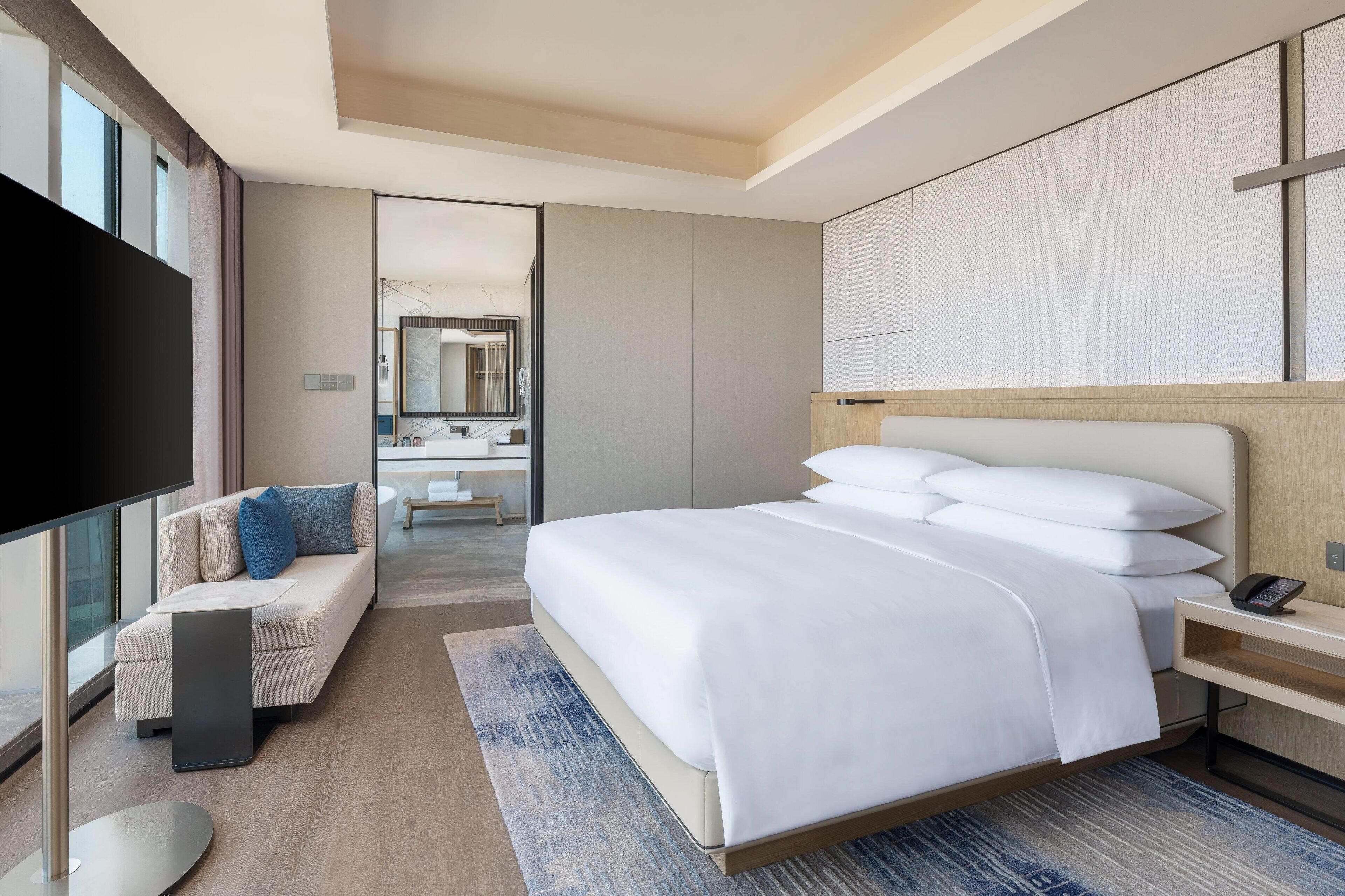 A modern hotel room with a large bed, a sofa, a TV, and a bathroom visible through an open door.