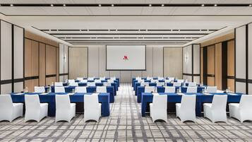 A conference room with rows of tables and chairs, a large screen at the front, and a patterned carpet.