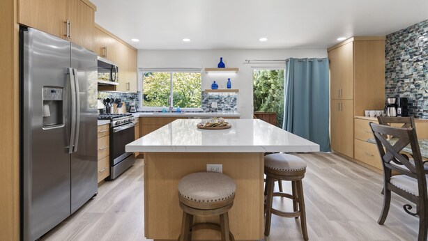 Een moderne keuken met houten kasten, roestvrijstalen apparatuur, een groot eiland met krukken en uitzicht op groen.