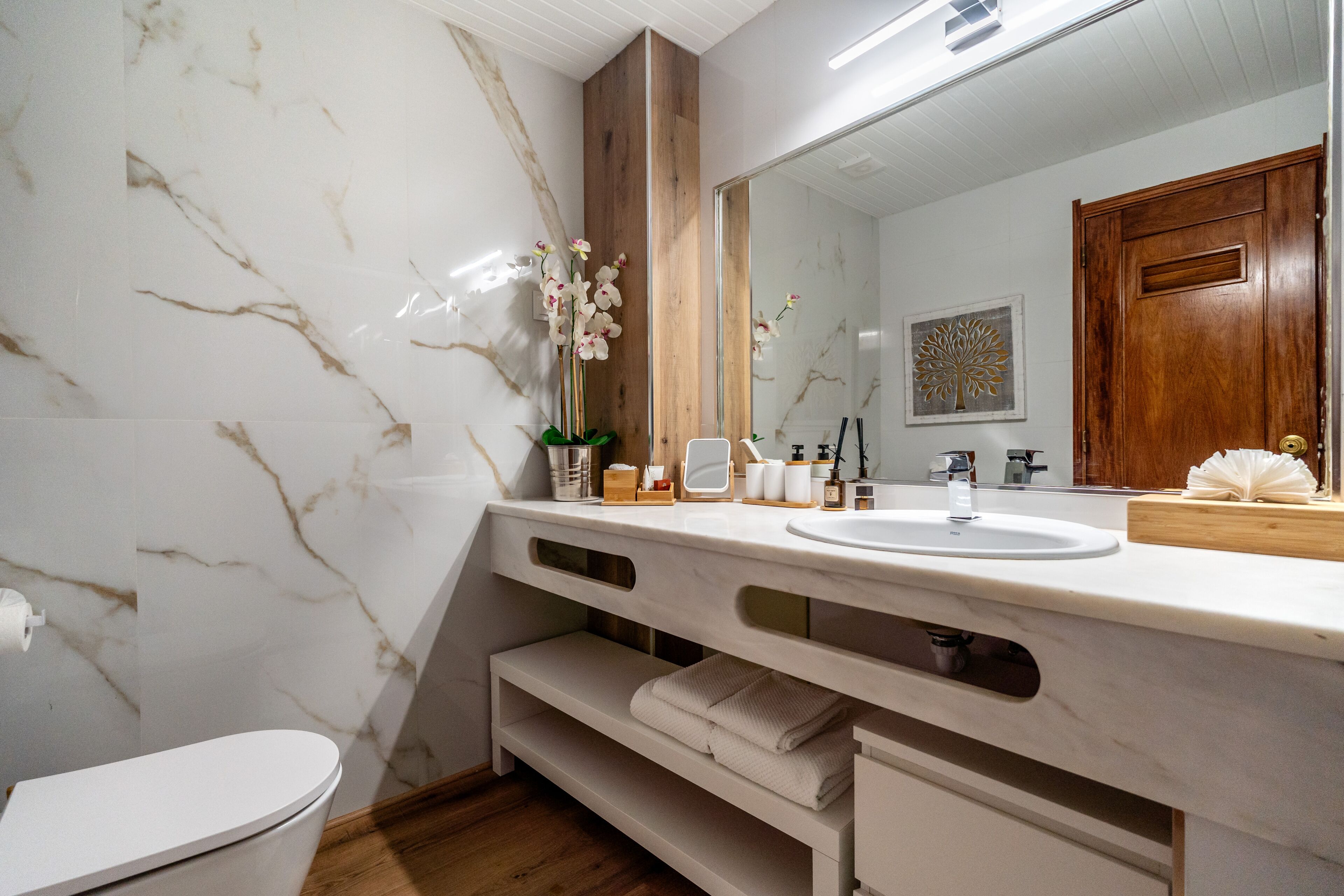 A bathroom with a marble wall, a wooden vanity, a sink, and a toilet.