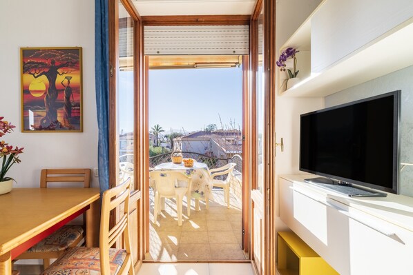 A balcony with a table and chairs, a TV on a white cabinet, and a painting on the wall.