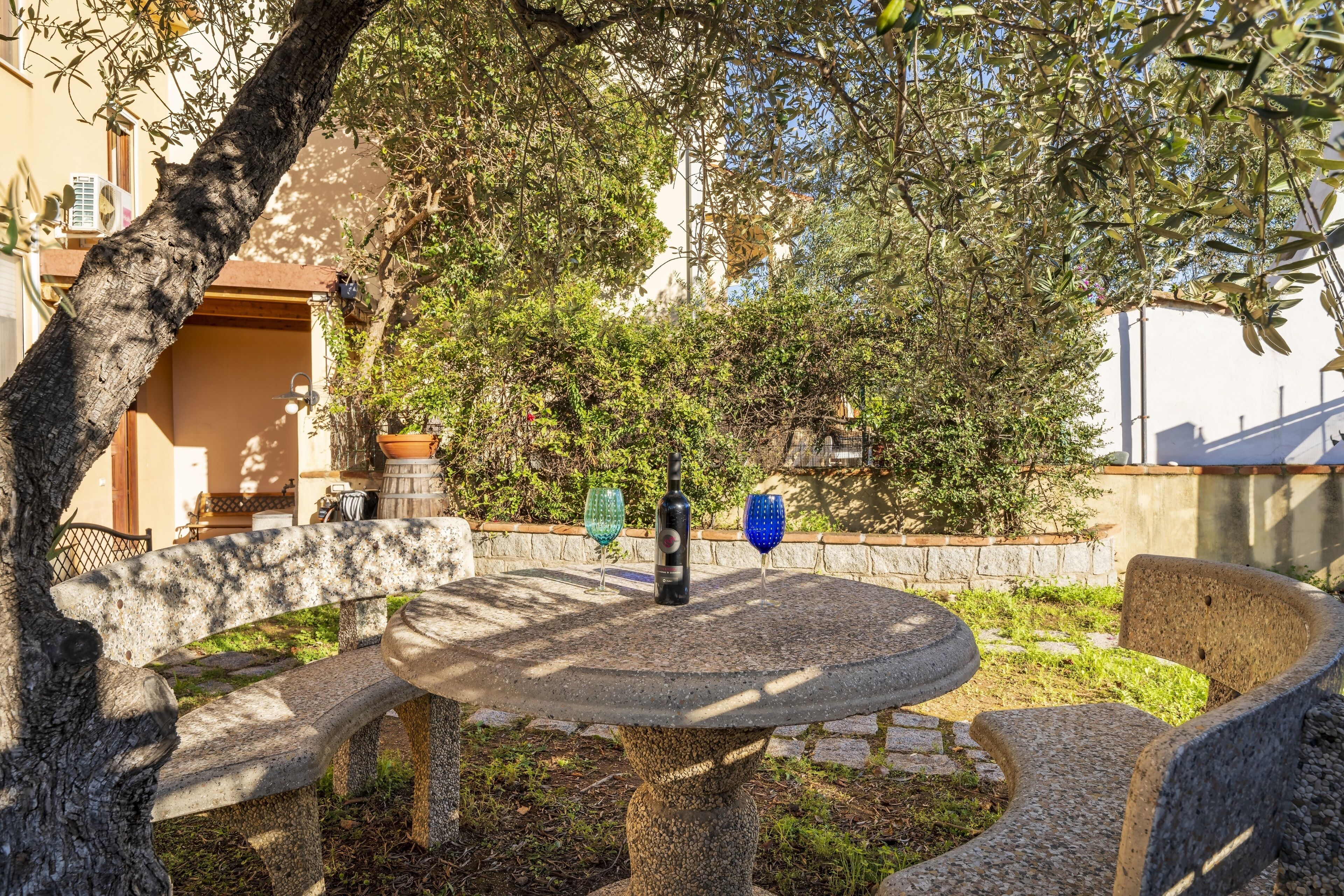 A stone bench with a table in a garden area, surrounded by trees and a building in the background.