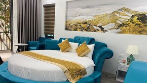 A round bed with a blue upholstered headboard and footboard, white bedding, and gold accents. A large landscape painting is on the wall behind the bed.