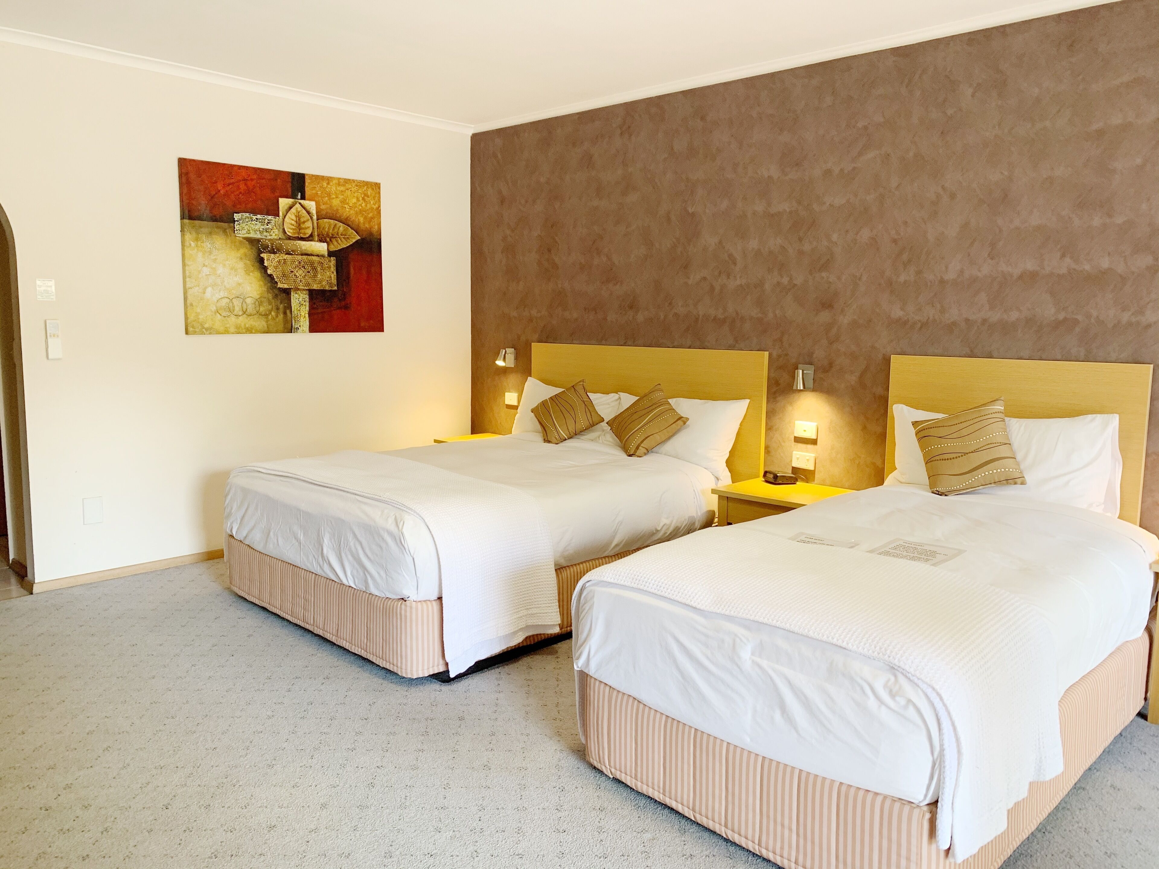 Habitación de hotel con dos camas, cada una con cabecero, mesitas de noche y lámparas. Hay un cuadro en la pared y piso alfombrado.
