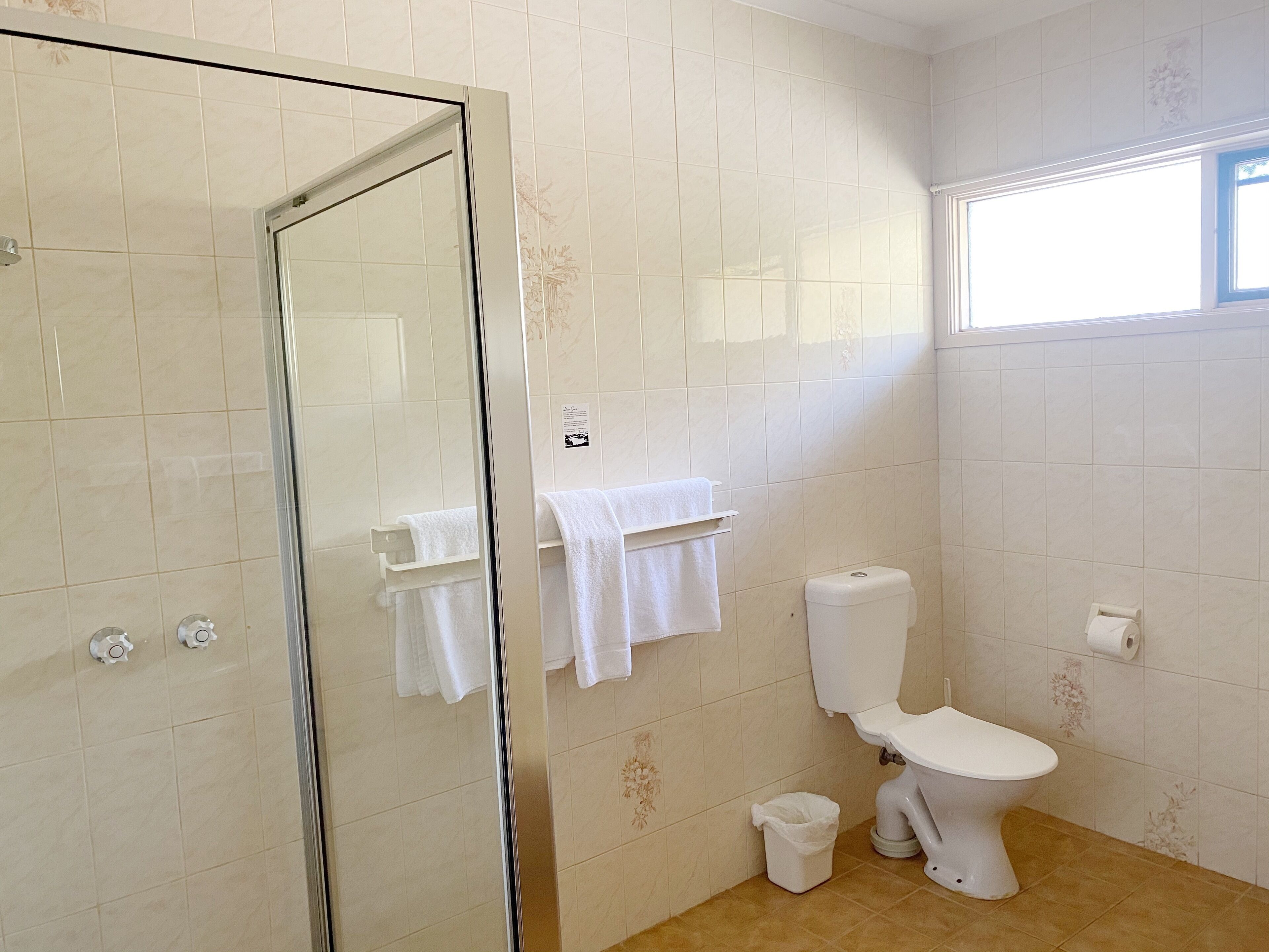 A bathroom with a glass shower door, a toilet, a small trash bin, and a window providing natural light.