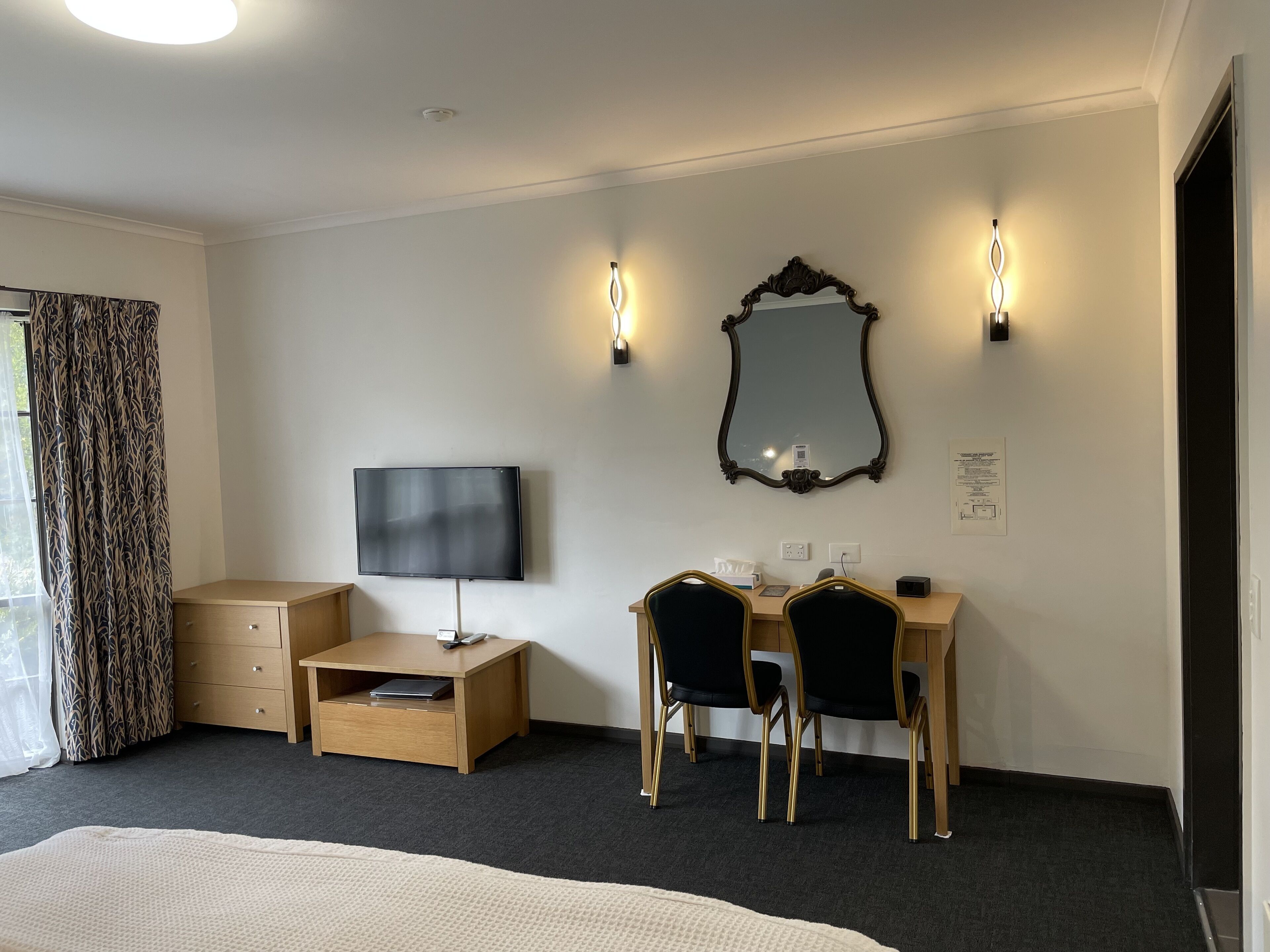 A hotel room with a bed, a desk with two chairs, a television, a mirror, and wall-mounted lights.
