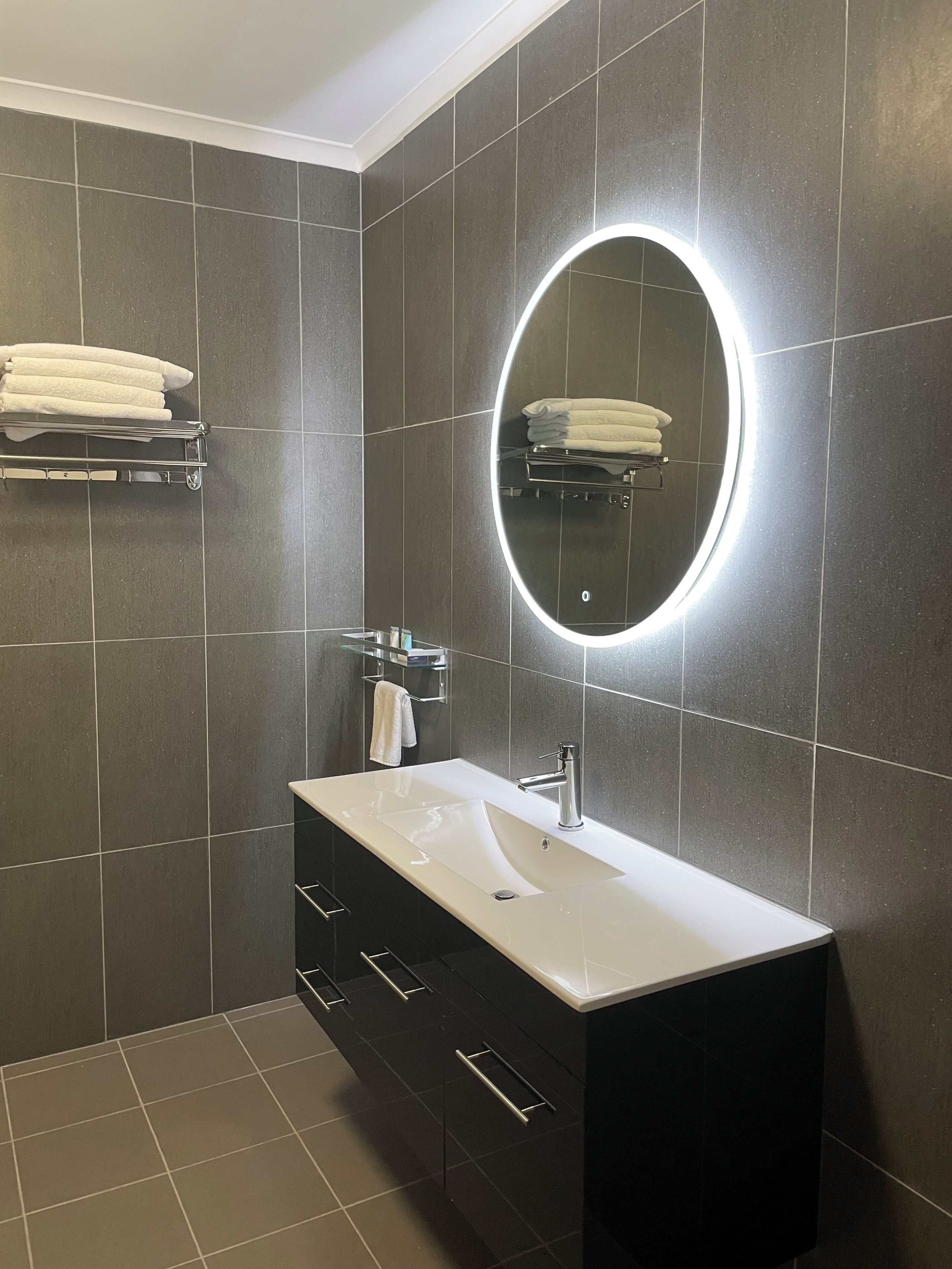 A modern bathroom with dark tiles, a white sink, a mirror with a light, and a towel rack.