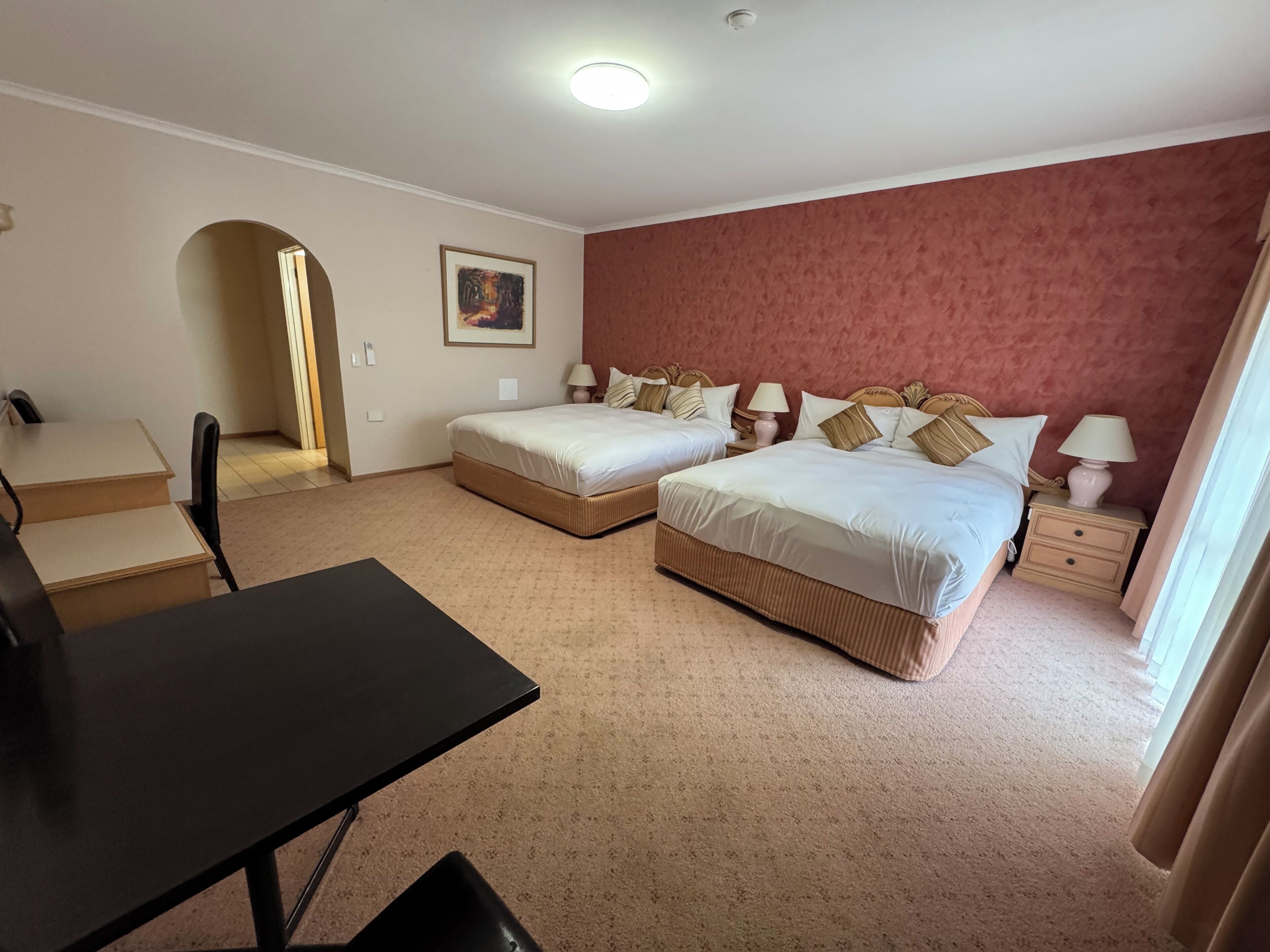 Een hotelkamer met twee bedden, een bureau en een stoel. Op de muur hangt een schilderij en er is een deur naar een andere kamer.