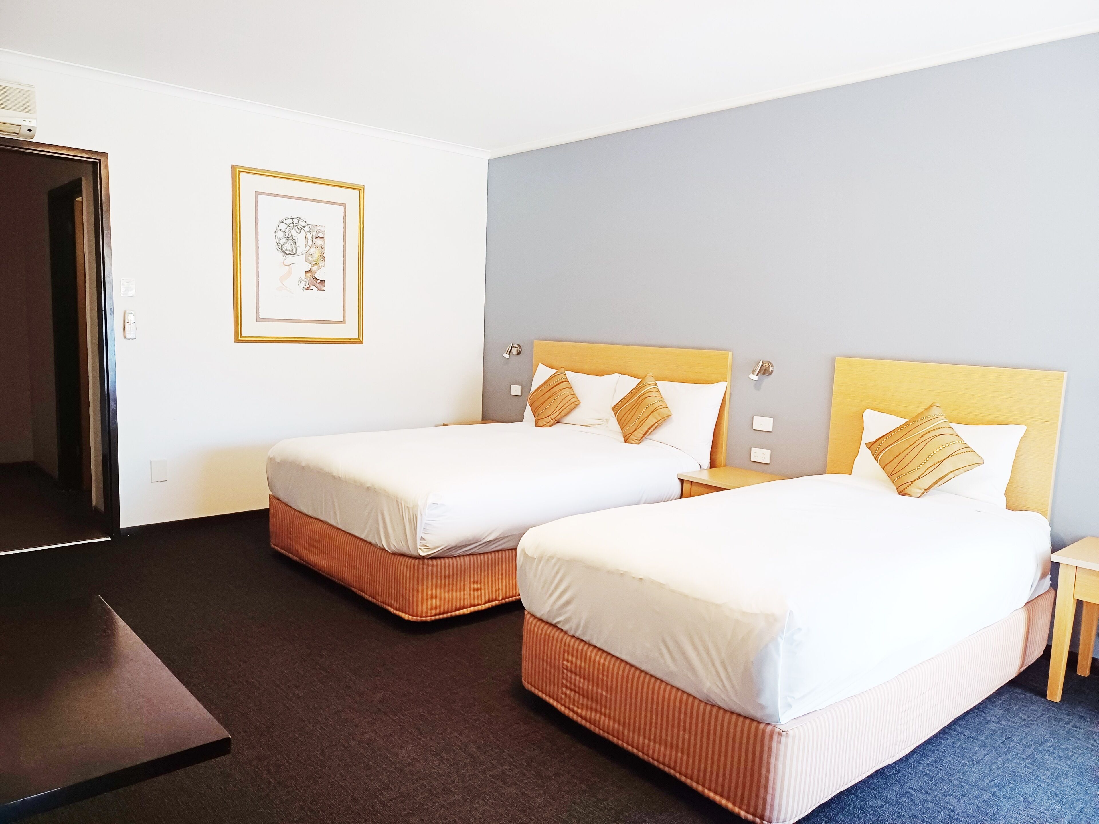 A hotel room with two single beds, a wooden headboard, a framed picture on the wall, and a small table.