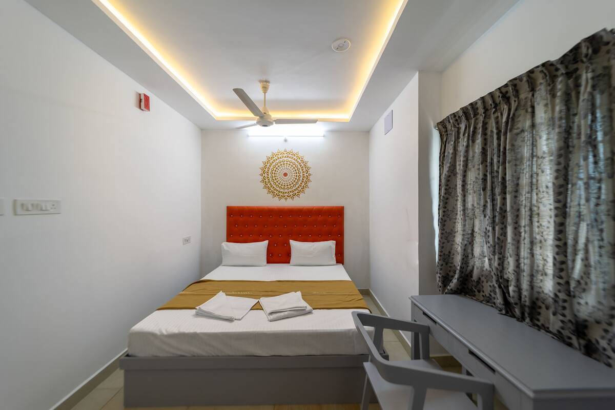 A hotel room with a bed, a ceiling fan, a decorative wall piece, a desk, and a window with curtains.