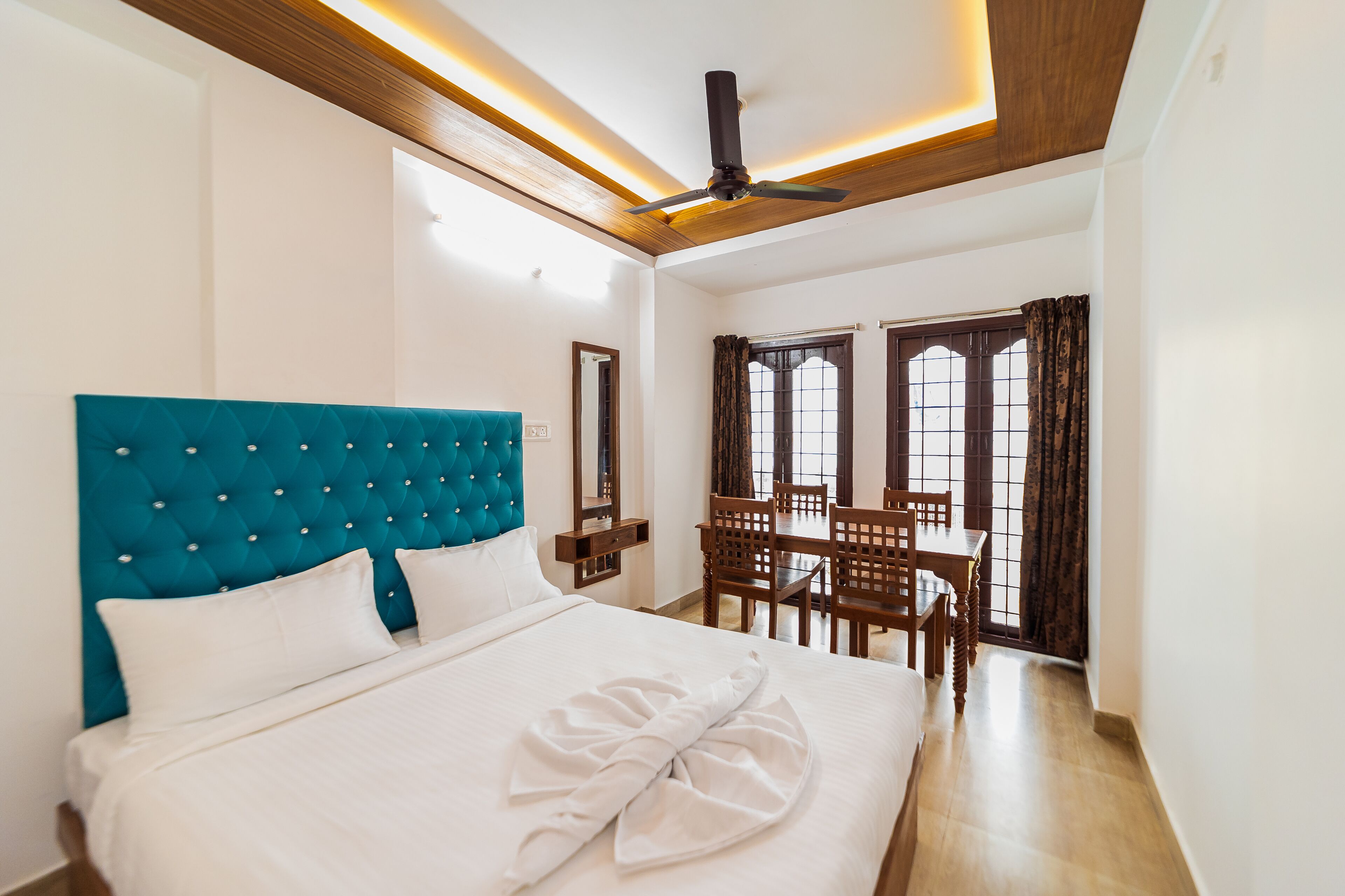 A hotel room with a large bed, a wooden ceiling, a dining area with a table and chairs, and a window with curtains.