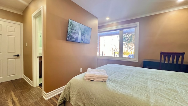 A bedroom with a bed, a television mounted on the wall, a window with a view of trees, and a door leading to a bathroom.