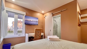A bedroom with a bed, a desk, a chair, a TV, a window, and a shower.