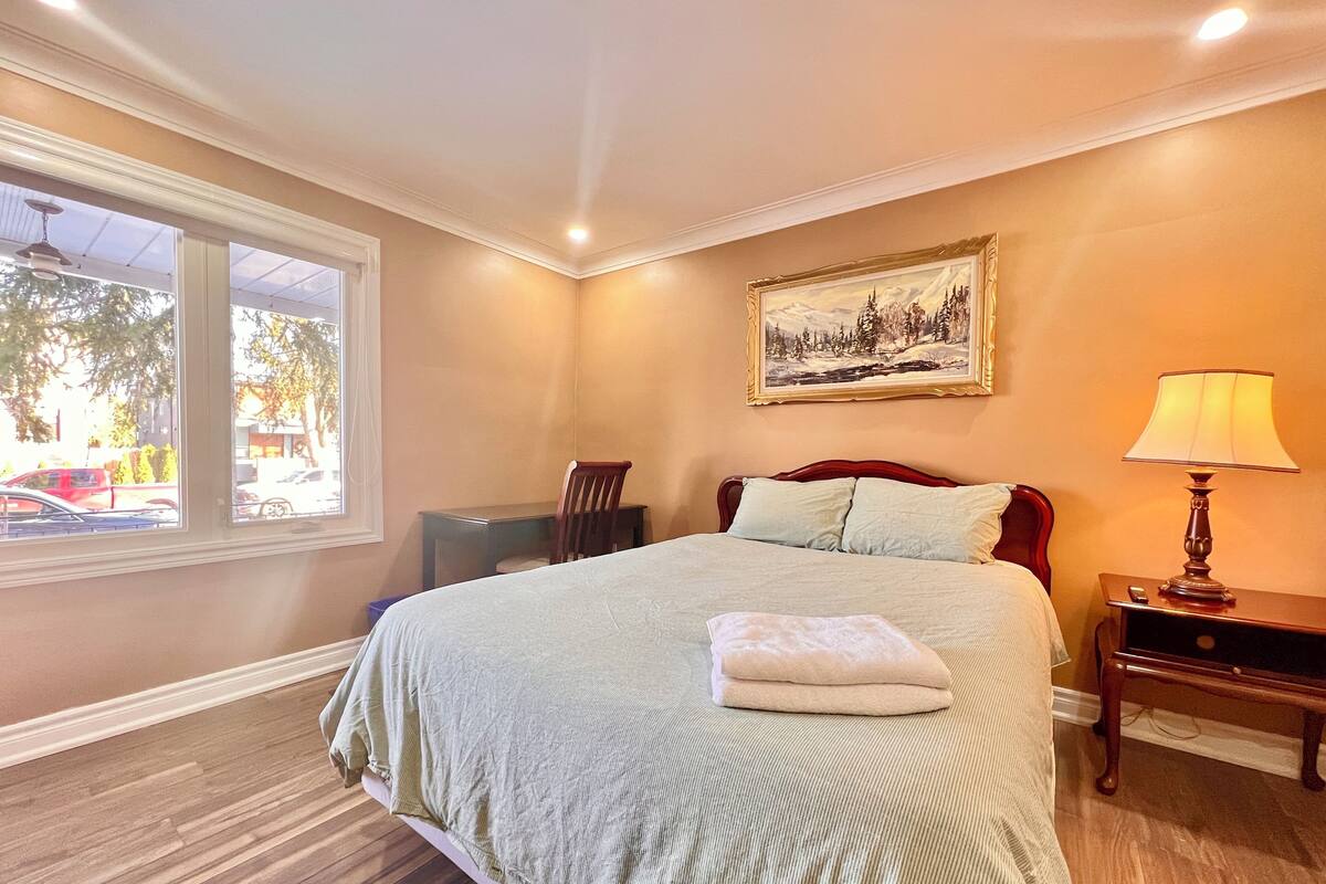 Un dormitorio con una cama grande, una mesita de noche, una lámpara, una silla, un cuadro y una ventana con vistas a la calle.