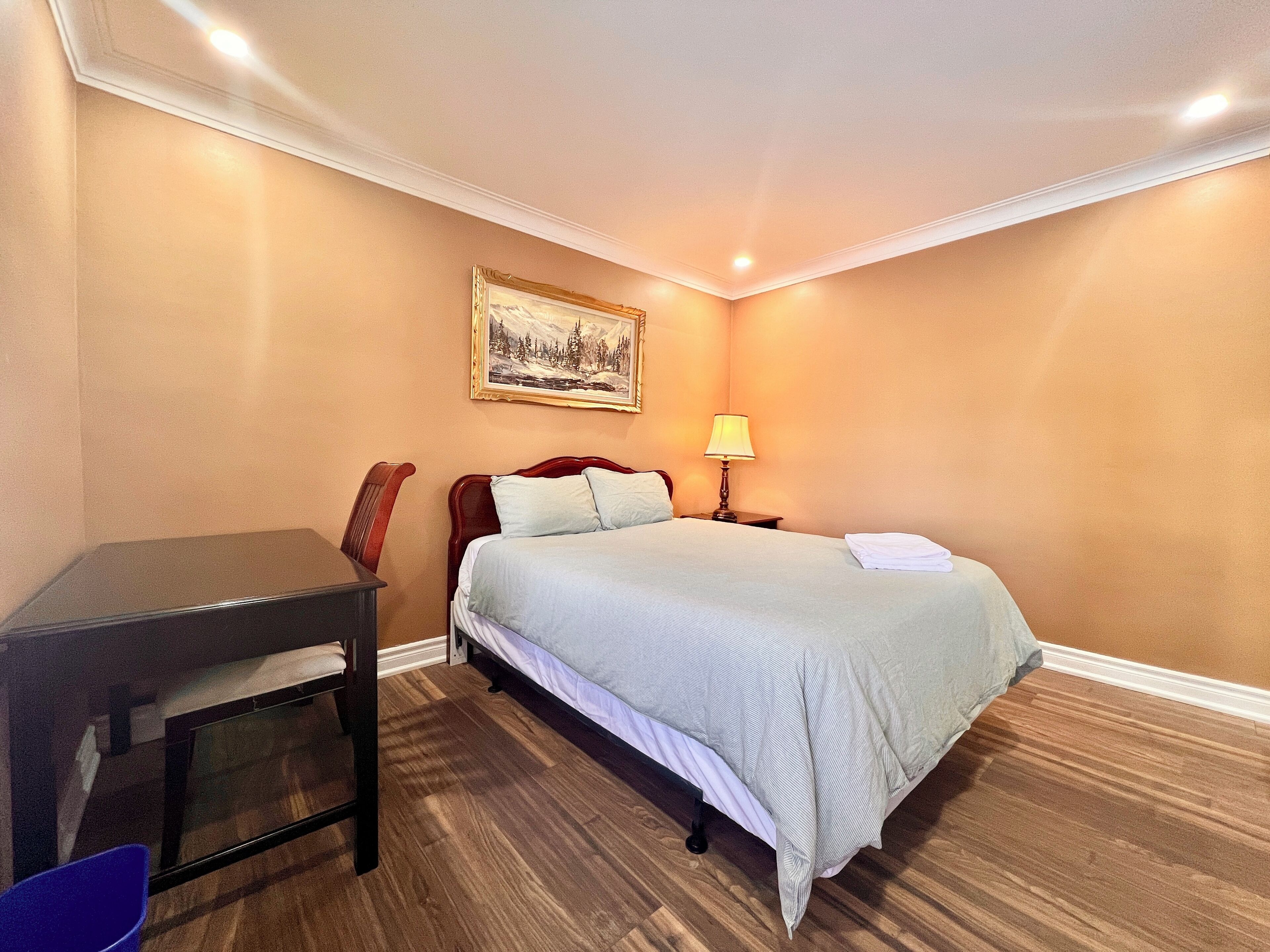 Quarto de hotel com cama, escrivaninha, cadeira, abajur e um quadro na parede.