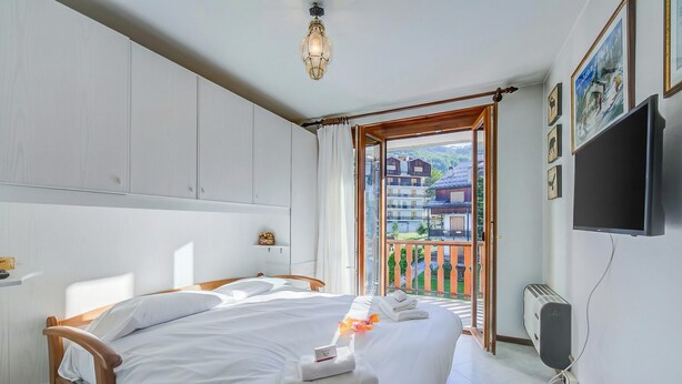 A hotel room with a bed, a TV, a window with a view of buildings, and a balcony.