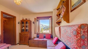 A cozy room with a wooden cabinet, a sofa with patterned cushions, a window with a mountain view, and a hanging light fixture.