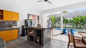 A modern kitchen with a built-in island, a dining area with a table and chairs, and a view of a backyard with a pool and plants. - Pasha Villa by Betterplace (Kerobokan)