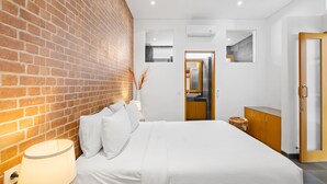A modern hotel room with a brick accent wall, a large bed, a bedside table with a lamp, and a view into the bathroom and kitchen. - Pasha Villa by Betterplace (Kerobokan)