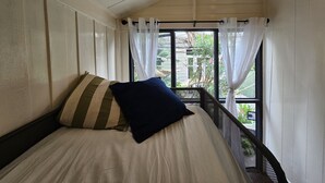 Una cama con un almohadón a rayas y uno oscuro, una ventana con cortinas de gasa, y vistas al exterior con vegetación.
