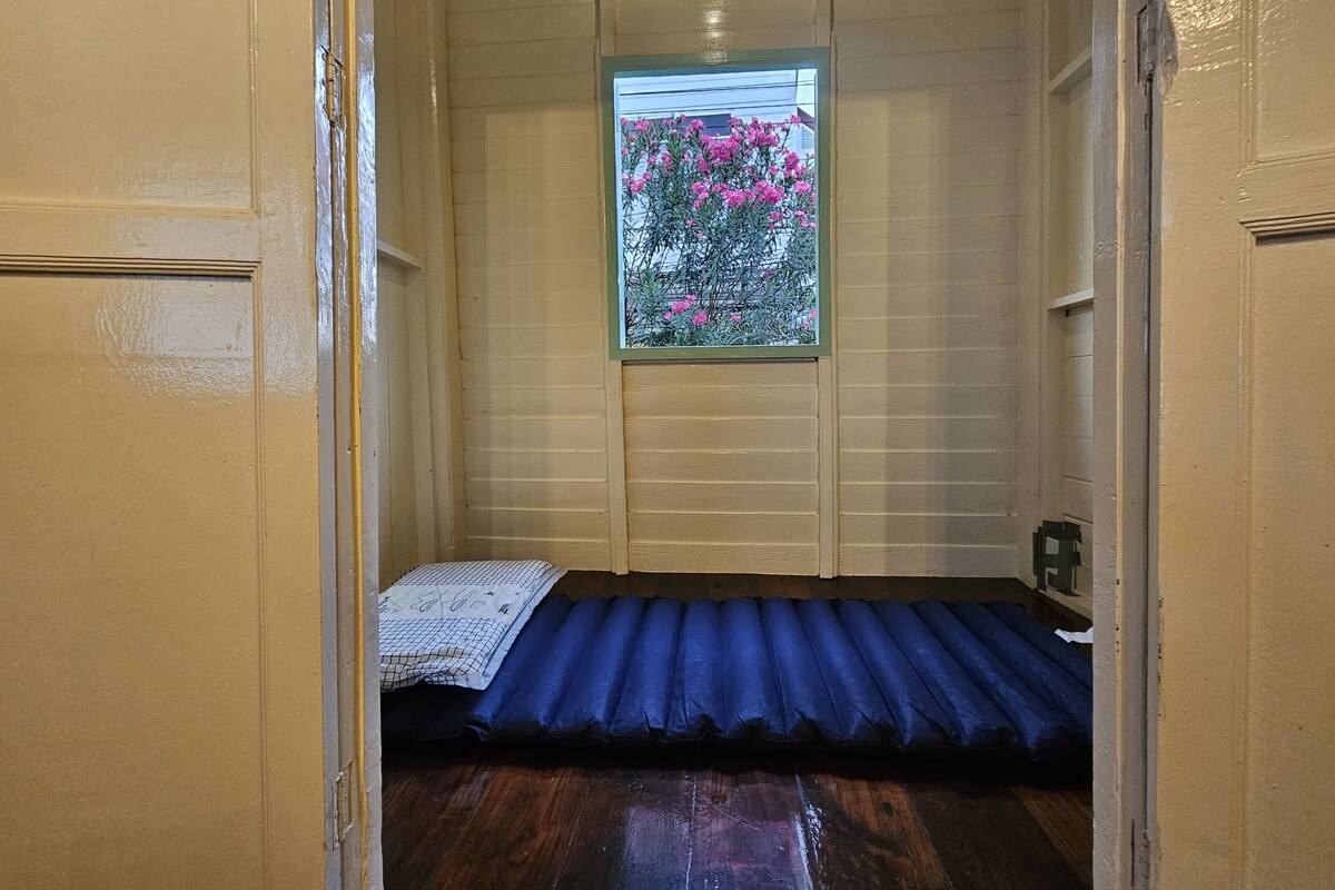 A small, enclosed room with a single bed, a window with a view of pink flowers, and wooden walls.