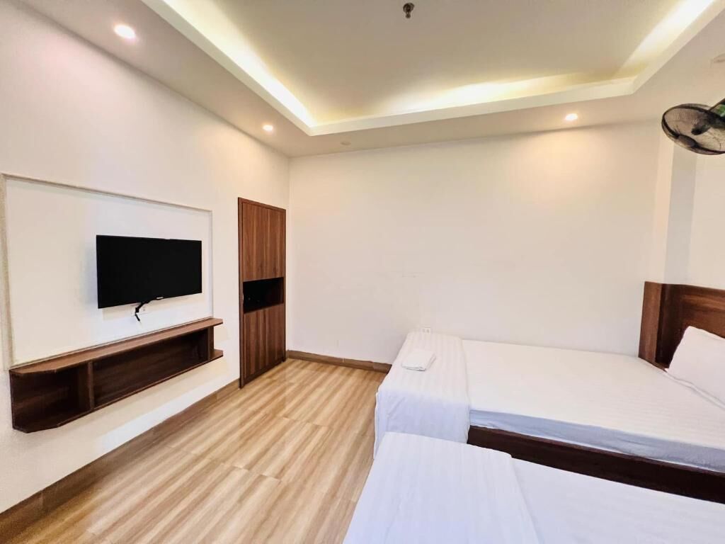 Ein Hotelzimmer mit Flachbildfernseher an der Wand, Holzboden, zwei Einzelbetten und Deckenventilator.