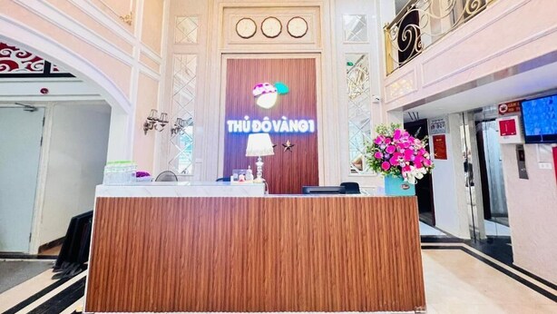 A reception desk with a wooden finish, a logo above it, and a decorative wall with a clock and a star. - Thu Do Vang Hotel Ha Dong By Bay Luxury (Hanoi)