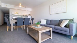 A modern living room with a grey sofa, a coffee table, a dining area with chairs, and a kitchen in the background. - Harbour View Apartment on 11th floor | King Bed (Darwin City)