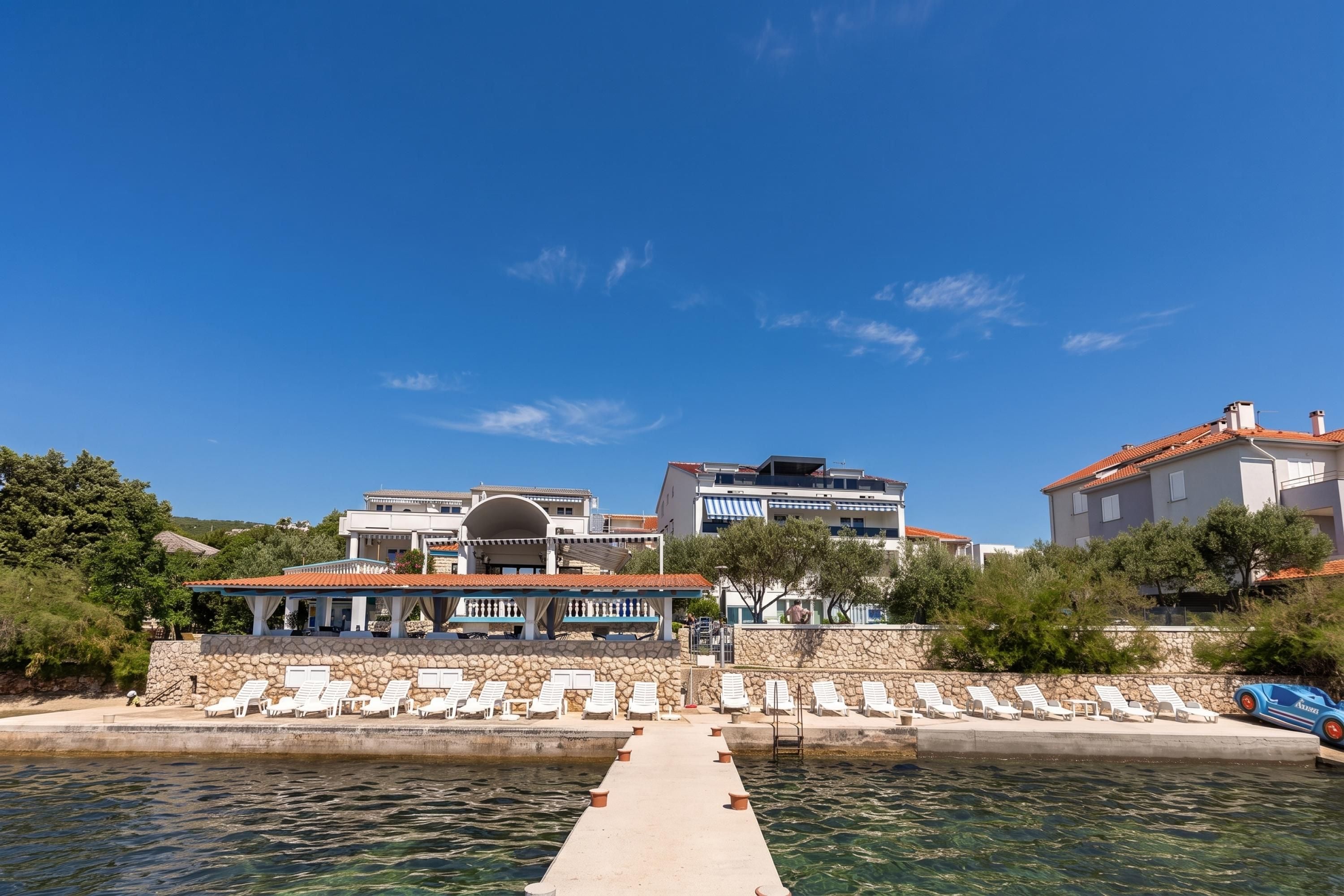 A waterfront property with a pool, lounge chairs, and a stone wall.