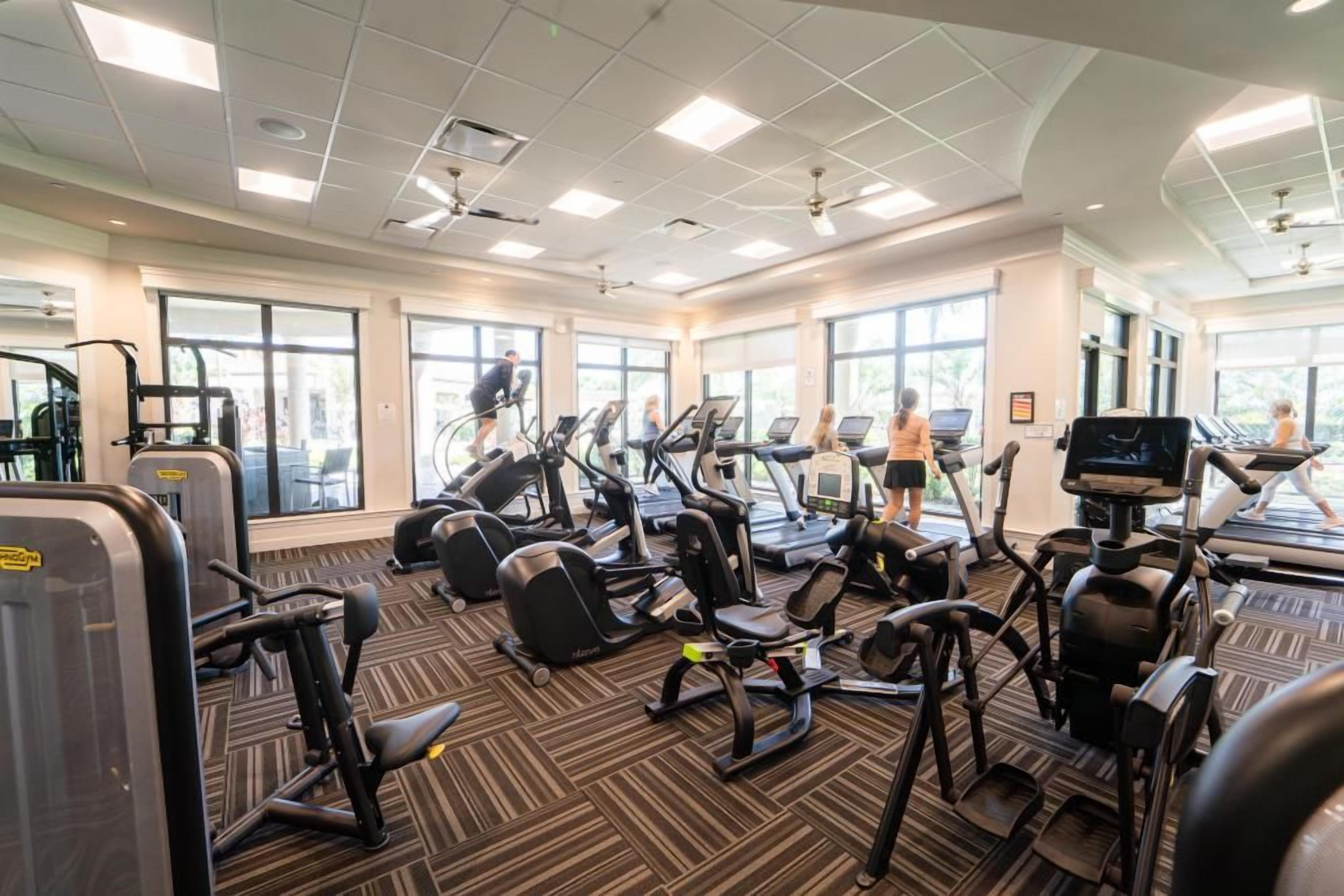 Ett gym med olika typer av träningsutrustning, stora fönster och ett golv täckt med matta.