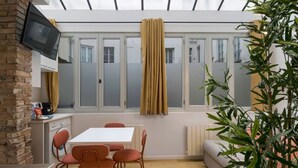 Une cuisine moderne avec un mur en briques, un téléviseur fixé au mur, et un coin repas avec une table blanche et des chaises rouges.
