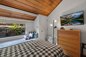 Een slaapkamer met een geruit beddegoed, een houten dressoir, een wandtv en een raam met uitzicht op groen en rotsen.