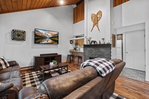 Een woonkamer met leren banken, een geruit vloerkleed, een open haard, een keukenhoekje en een kunstwerk aan de muur.
