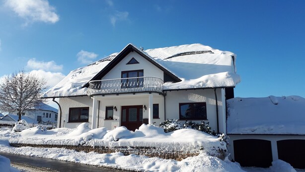 Une maison recouverte de neige, dotée d’un balcon et d’un mur en pierre, sous un ciel bleu clair.