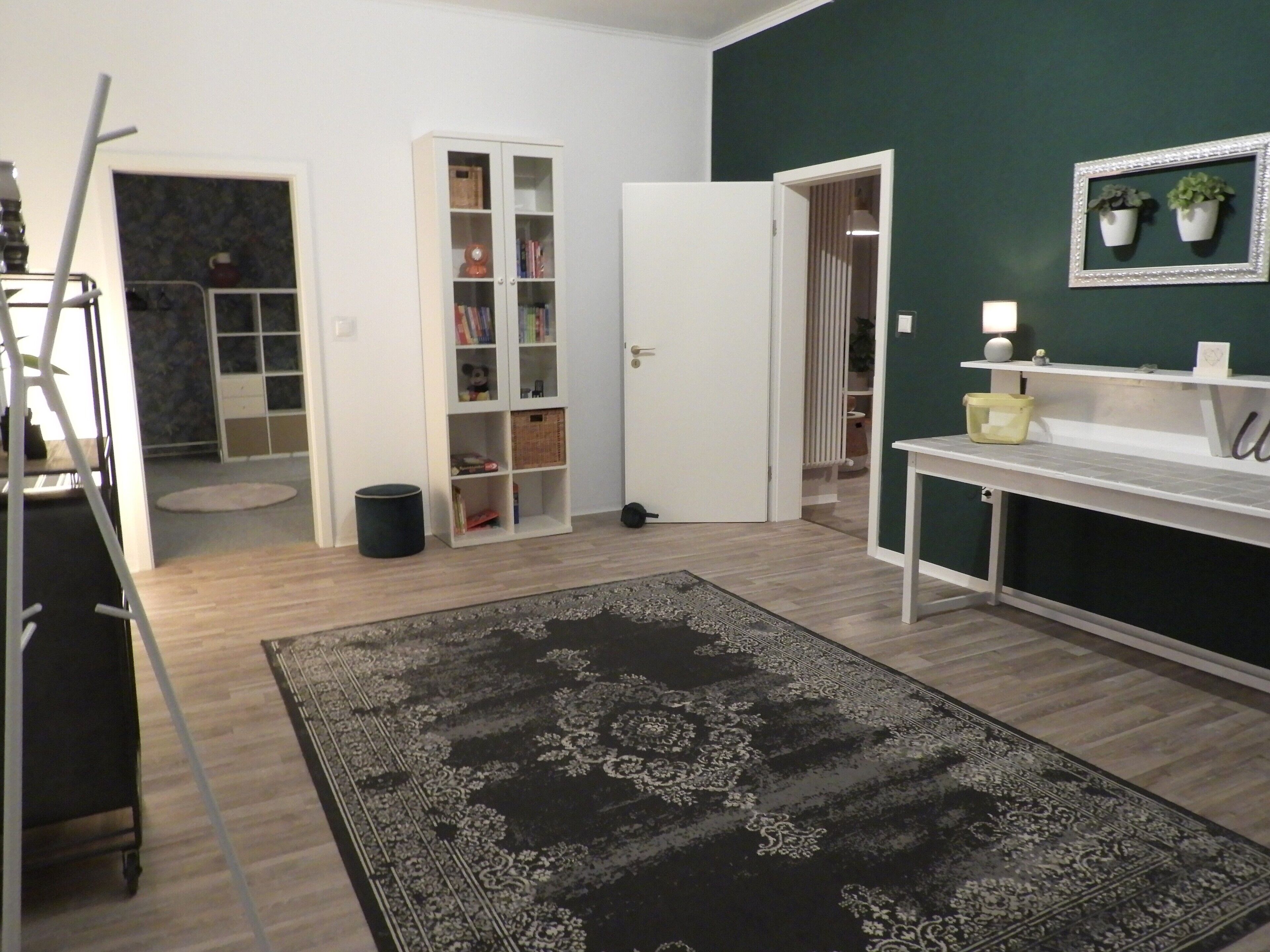 Une pièce avec un plancher en bois, un tapis noir à motifs, un bureau blanc, une étagère blanche ornée de plantes, une porte blanche et une bibliothèque.