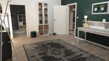 Une pièce avec un plancher en bois, un tapis noir à motifs, un bureau blanc, une étagère blanche ornée de plantes, une porte blanche et une bibliothèque.